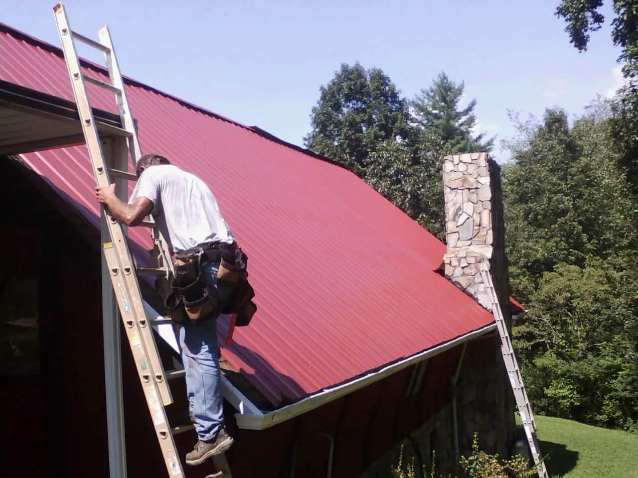 Roofer on a ladder beside a red metal roof and stone chimney — Soffit & Fascia Repair by Roofing Brothers in Henderson
